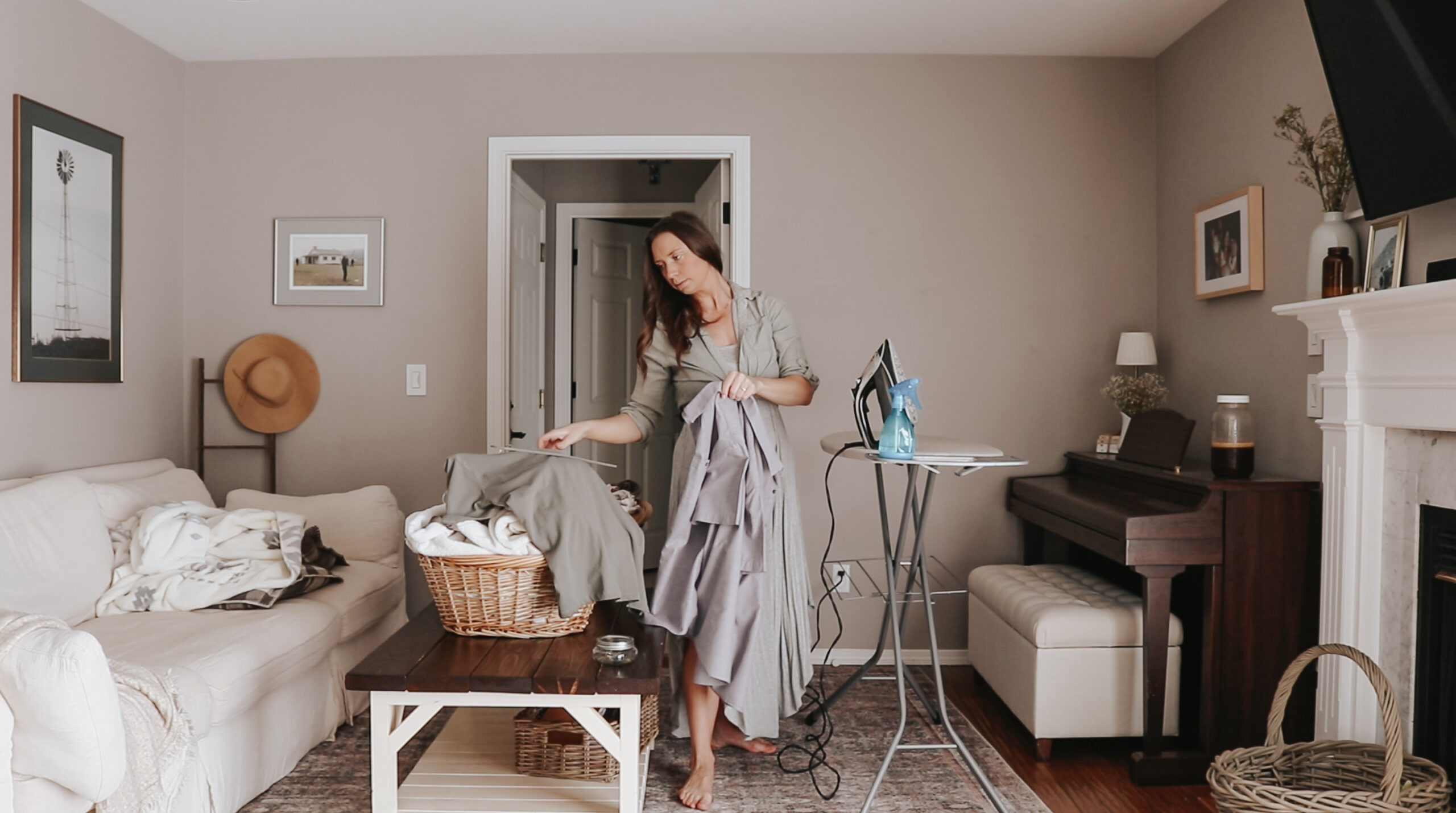 Christian homemaker does laundry and ironing in her living room, she is holding a shirt standing between a coffee table with a laundry basket and an ironing board
